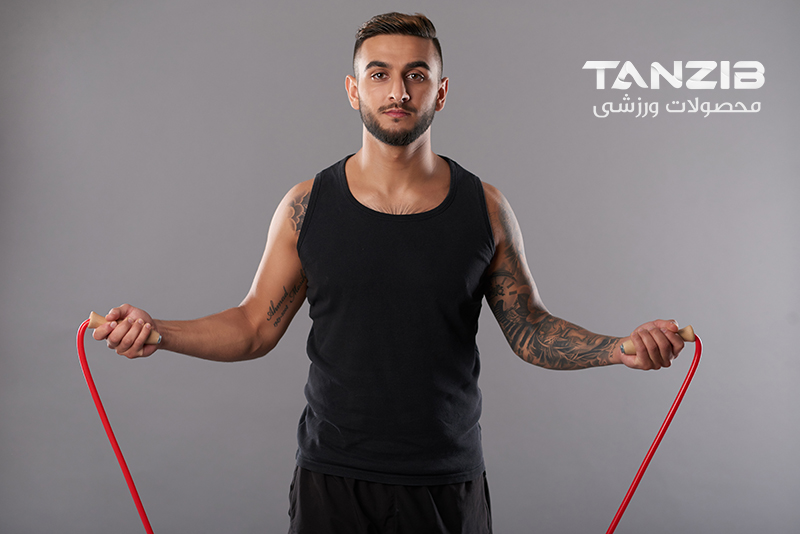 Handsome tattooed man in black sportswear looking seriously at camera while jumping rope on gray background