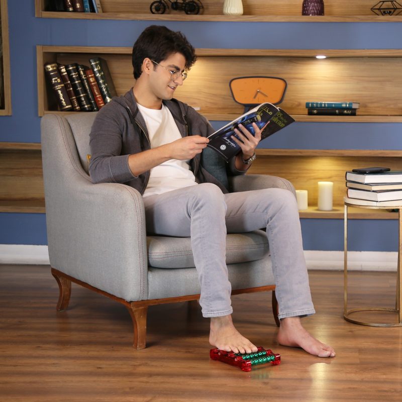 پسر جوانی در حال استفاده از ماساژور کف پای مهره ای در حالت نشسته و در حال خواندن مجله رو مبل در خانه