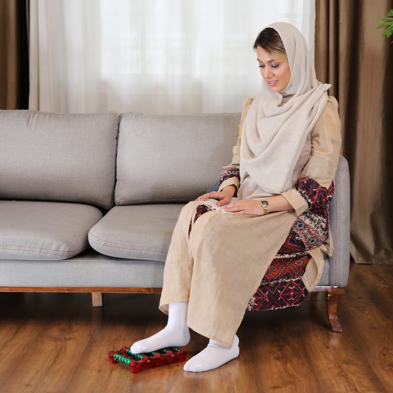 خانم جوانی در حال استفاده از ماساژور کف پای مهره ای در حالت نشسته روی مبل در فضای خانه