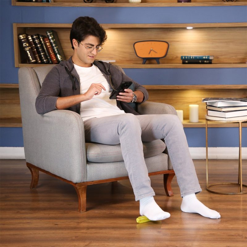 فردی در حال استفاده از ماساژور کف پا تن‌زیب مدل رولی روی مبل در فضای خانه در حال کار با گوشی