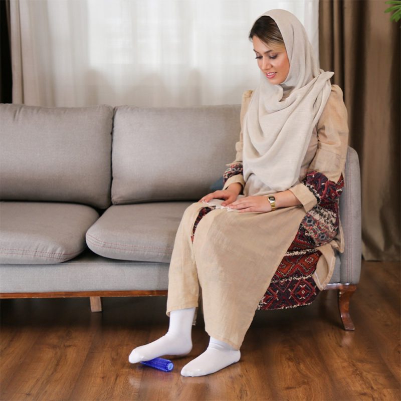 خانم جوانی در حال استفاده از ماساژور کف پا تن‌زیب مدل رولی آبی روی مبل در فضای خانه
